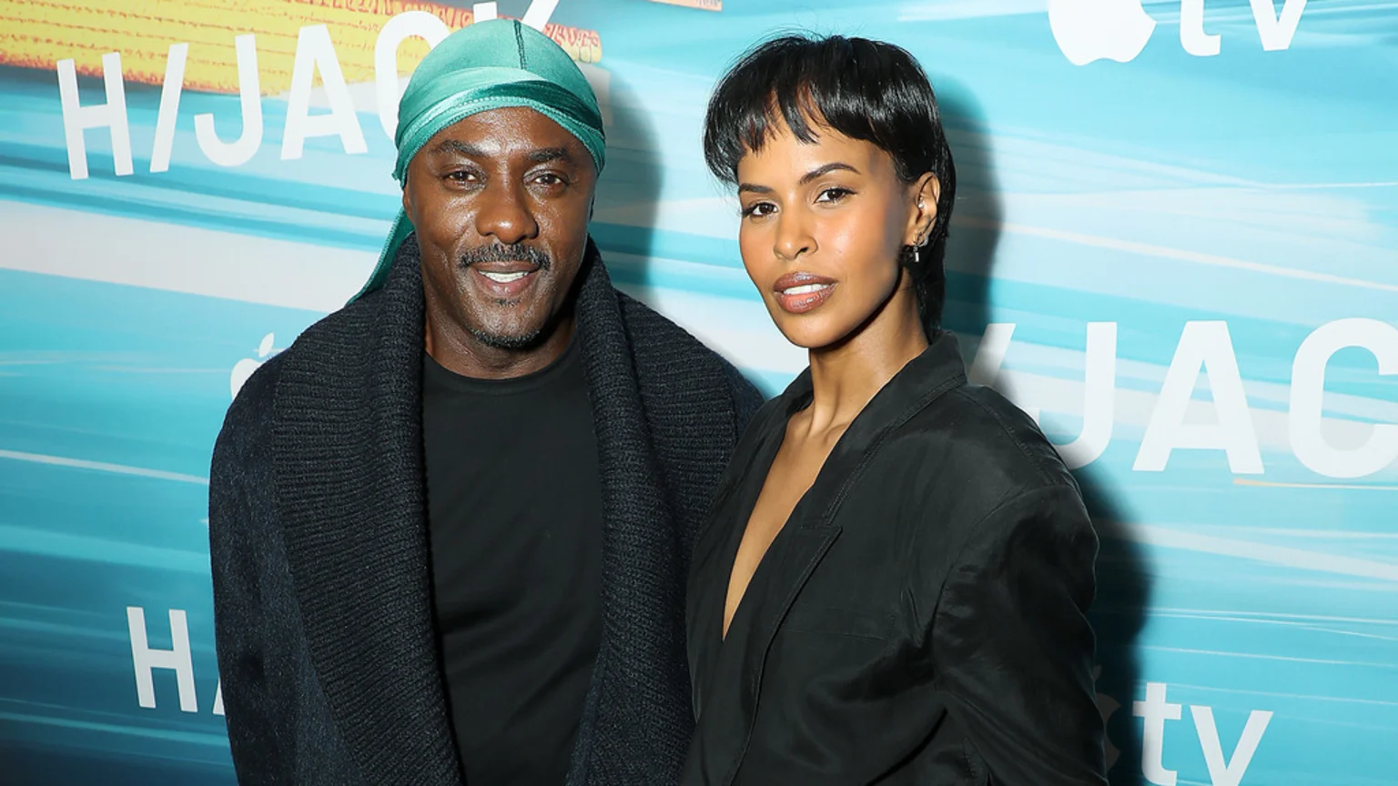 Idris Elba, wearing a teal durag and dark cardigan, smiles next to Sabrina Elba, who wears a black top, in front of a blue "HIJACK" and "Apple TV" backdrop.