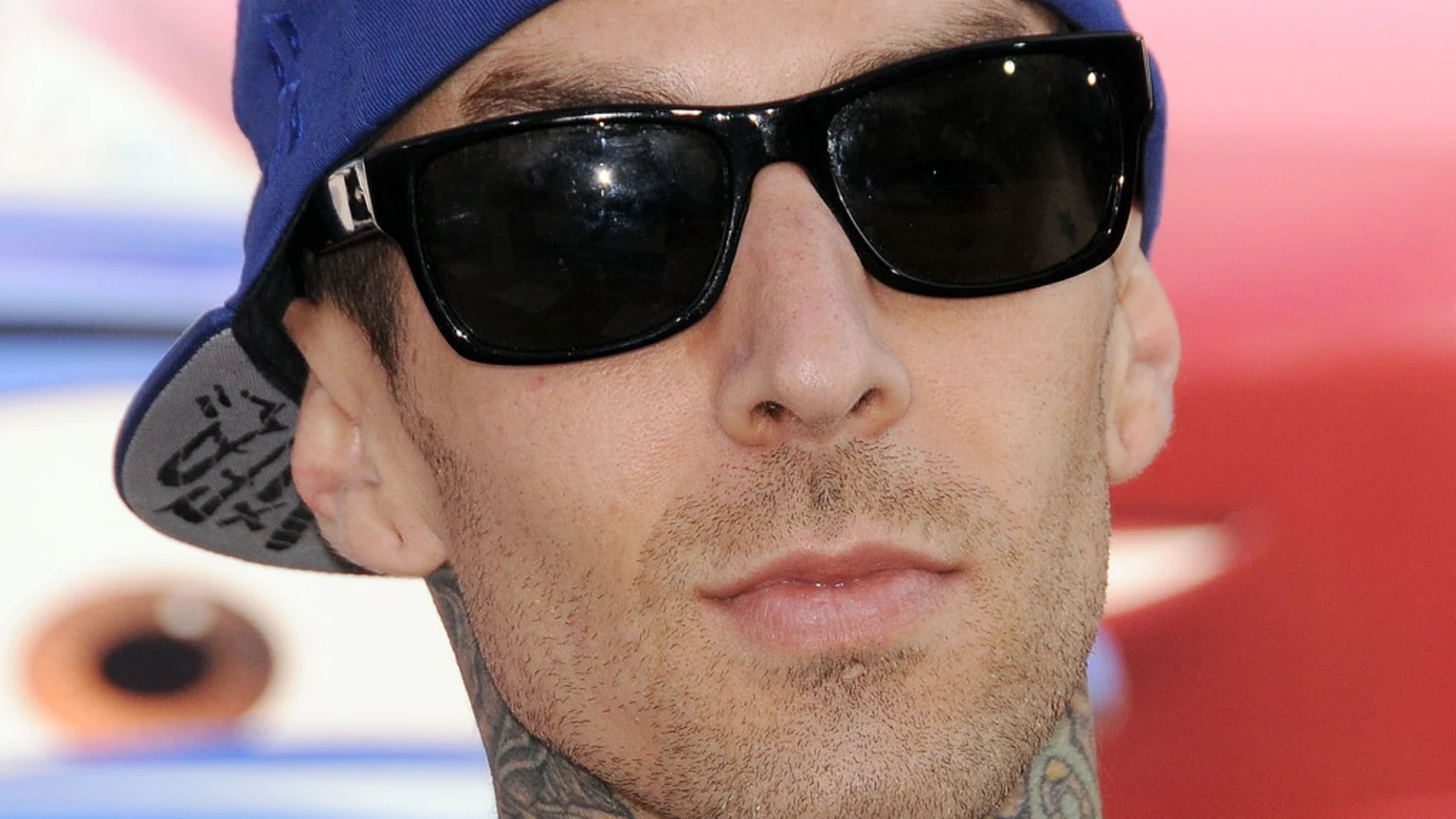 Close-up of Travis Barker wearing black sunglasses and a blue baseball cap backward, with stubble and neck tattoos visible.