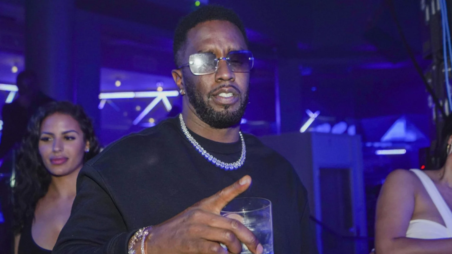 Sean 'Diddy' Combs wearing sunglasses and diamond jewelry, holding a drink and gesturing in a blue-lit club.