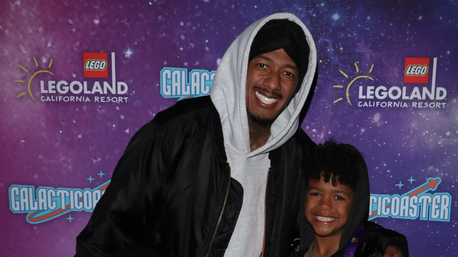 A smiling Nick Cannon in a black turban and grey hoodie poses with a young, smiling child against a purple Legoland California Resort backdrop.
