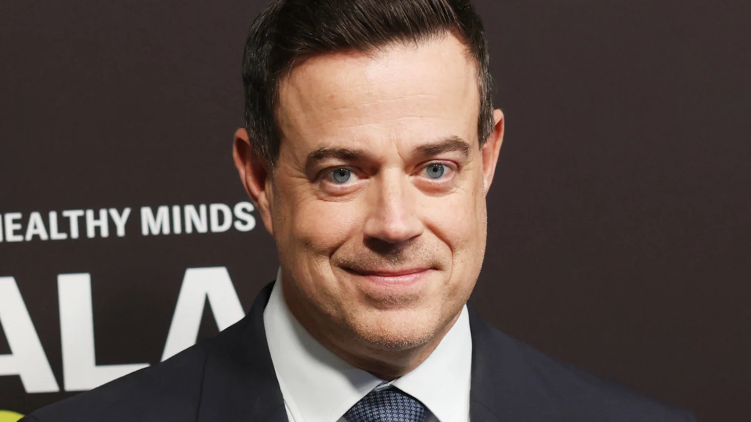 Carson Daly, a smiling middle-aged man with blue eyes and dark hair, wears a dark suit, white shirt, and patterned tie against a dark background with partially visible white text.