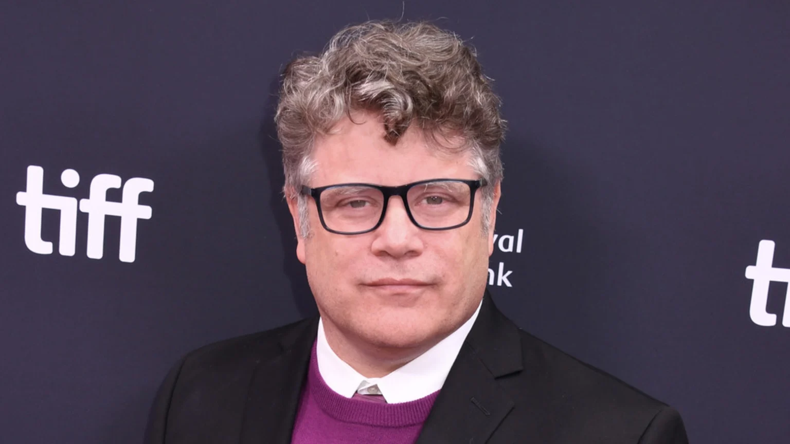 Actor Sean Astin, with curly gray hair and glasses, wears a black suit jacket and purple sweater, posing against a dark background with the white "tiff" logo visible.