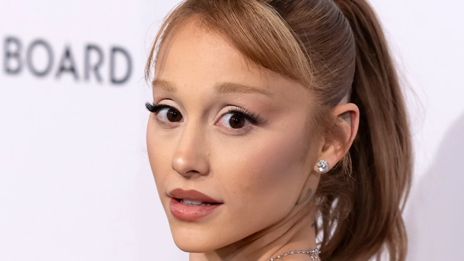 Close-up of Ariana Grande with light brown hair, wearing prominent eyelashes, a sparkling earring, and a necklace, looking wide-eyed with a slight smile; a blurry 'BOARD' sign is visible.