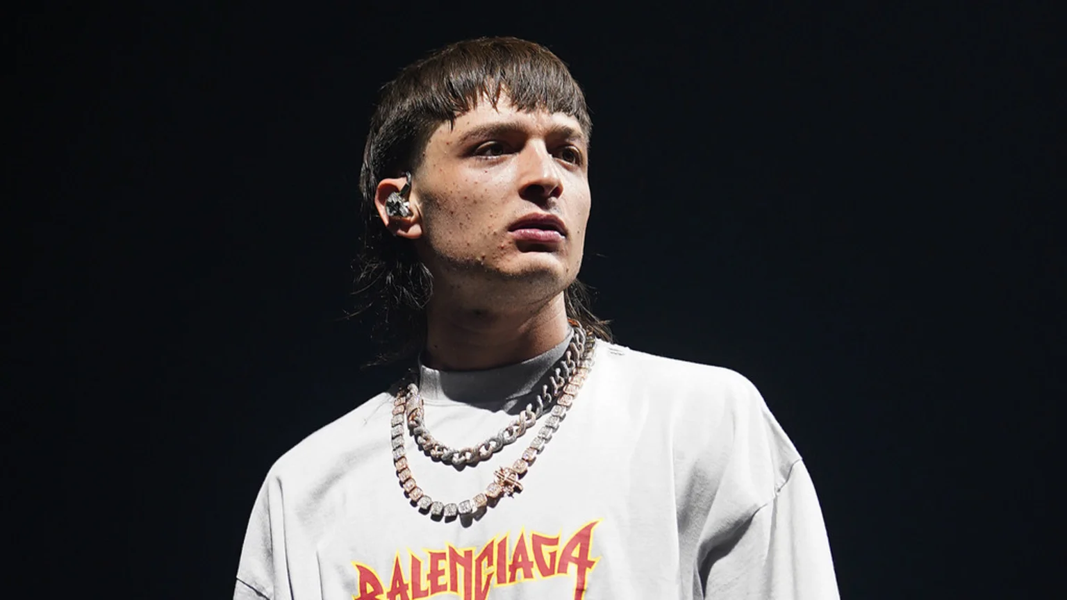 A man with a mullet haircut and an earpiece, wearing two layered chains and a light t-shirt with "BALENCIAGA," looks slightly up and to the right against a dark background.