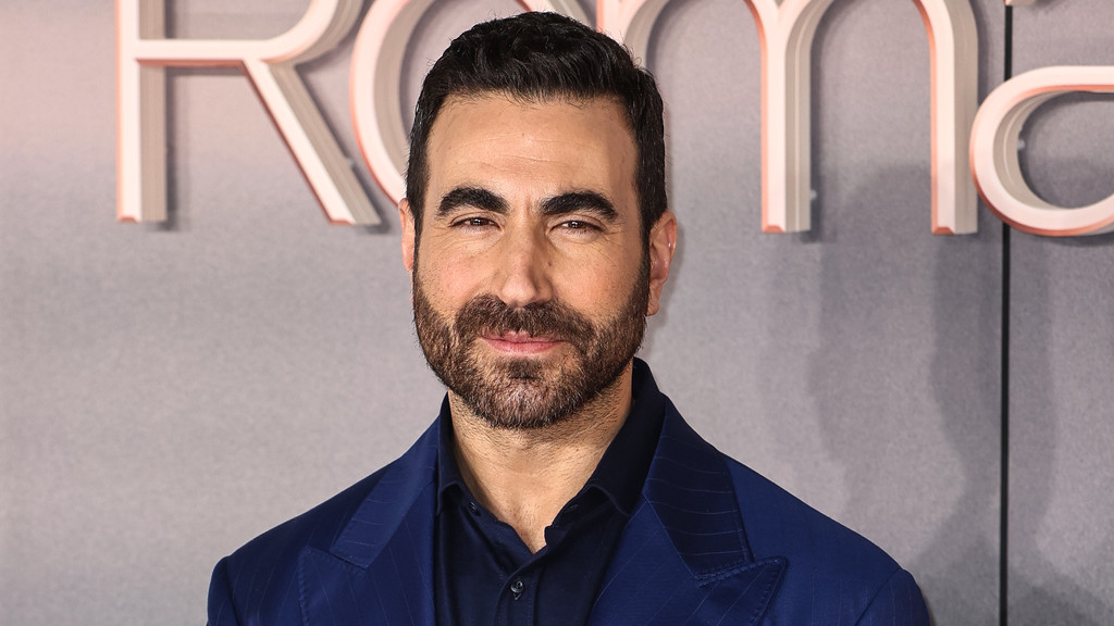 Brett Goldstein, a man with dark hair and a beard, smiles slightly while wearing a dark blue pinstripe suit jacket and dark blue shirt against a gray background with light letters.