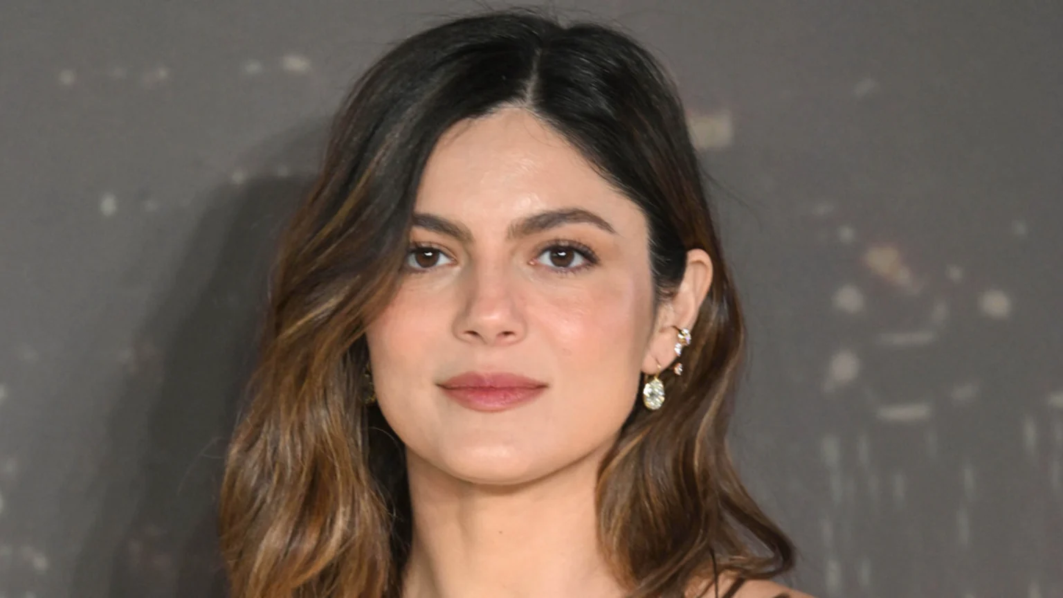 A headshot of actress Monica Barbaro with brown, wavy hair and natural makeup, looking directly forward. She wears an ear cuff and a drop earring.