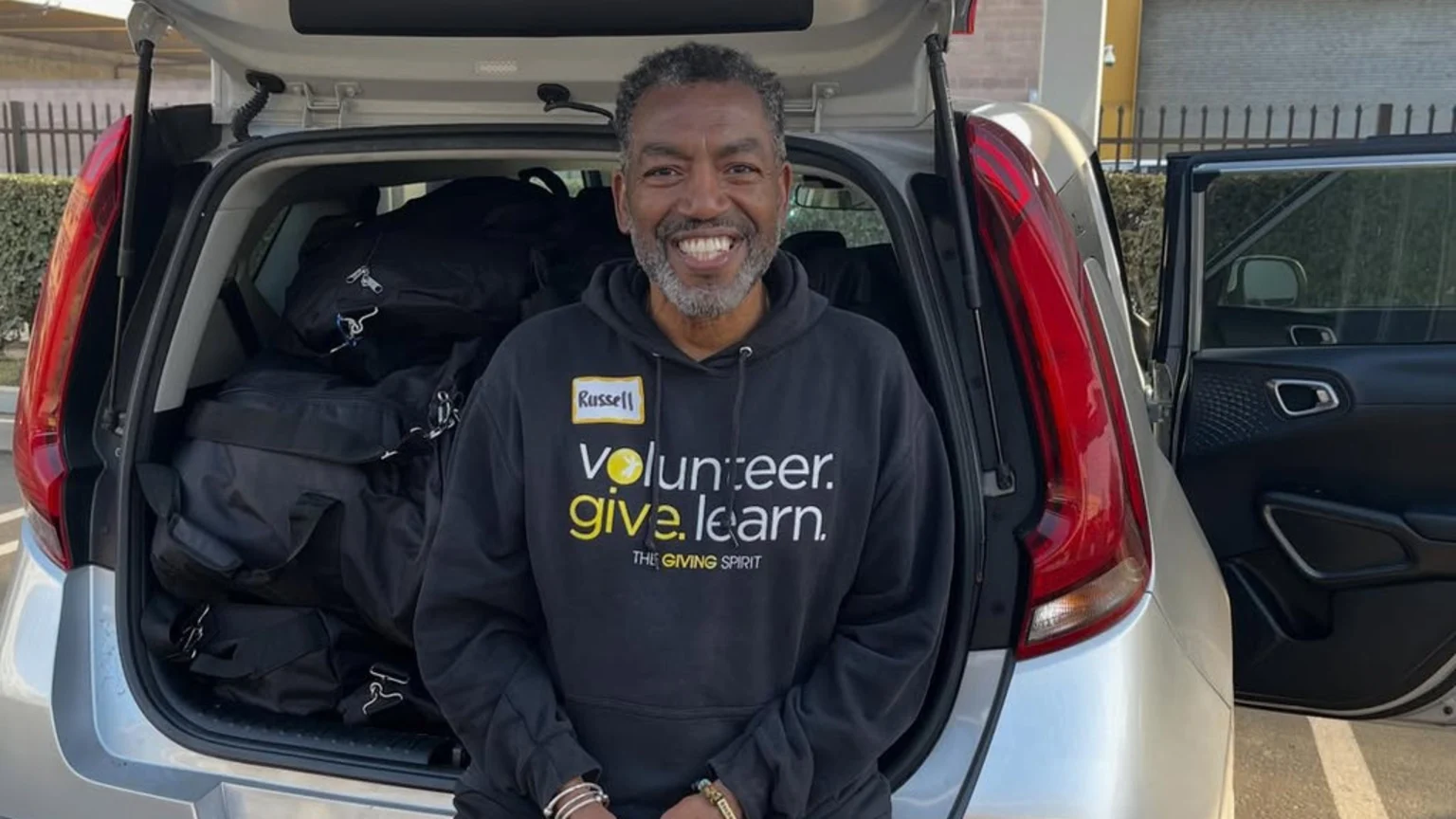 A smiling man with a name tag reading "Russell" wears a black "volunteer. give. learn." hoodie and stands in front of an open car trunk filled with black bags.