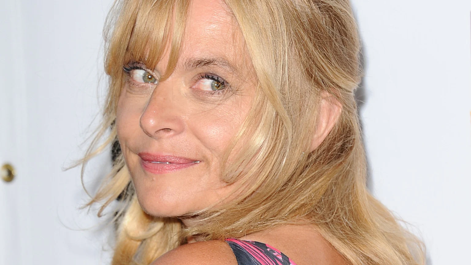A close-up of a blonde woman with light eyes looking directly over her shoulder at the viewer, smiling softly.