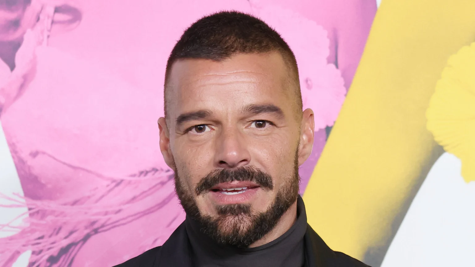 Close-up of Ricky Martin smiling slightly, with short dark hair and a beard, wearing a black turtleneck against a blurred pink and yellow background.