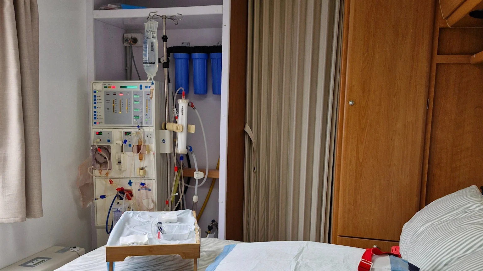 A medical machine, likely a dialysis unit, with an IV drip, tubing, and three blue water filters, stands next to a bed with medical supplies in a patient room.