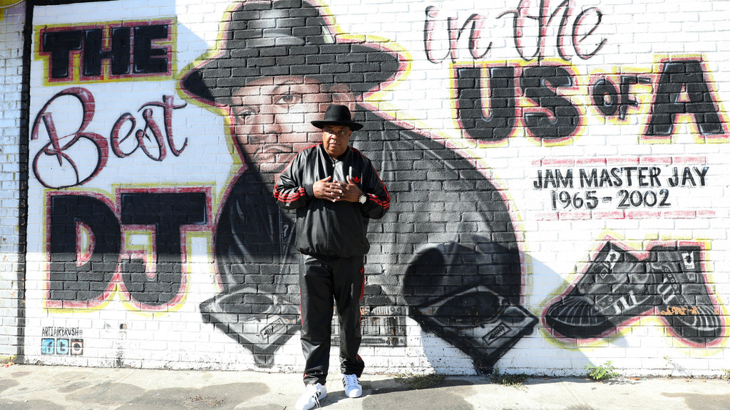 A man in a black tracksuit stands before a brick wall mural featuring a portrait of Jam Master Jay, turntables, and Adidas sneakers, with text: "THE Best DJ in the US OF A" and "JAM MASTER JAY 1965-2002".