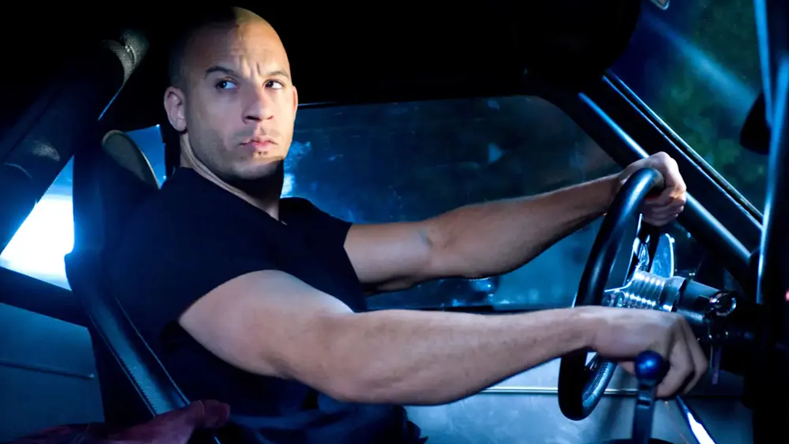 Close-up of actor Vin Diesel smiling, wearing aviator-style sunglasses, against a dark background.