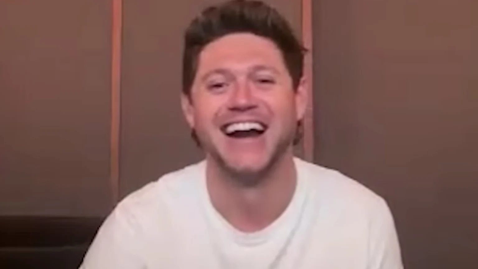 A man with light brown hair and a white t-shirt laughs broadly, revealing his teeth, against a reddish-brown wall.