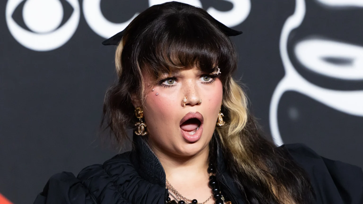 A woman with dark hair, blonde streaks, bangs, and facial piercings looks surprised with her mouth open, wearing a black top, layered necklaces, and Chanel earrings against a dark background with blurry white shapes.