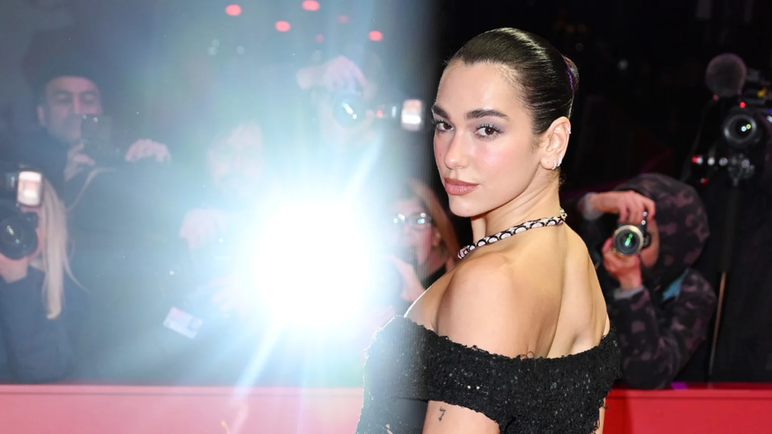 Dua Lipa, wearing a black off-the-shoulder dress and a necklace, looks over her shoulder at the viewer as bright camera flashes illuminate the scene with photographers in the background.