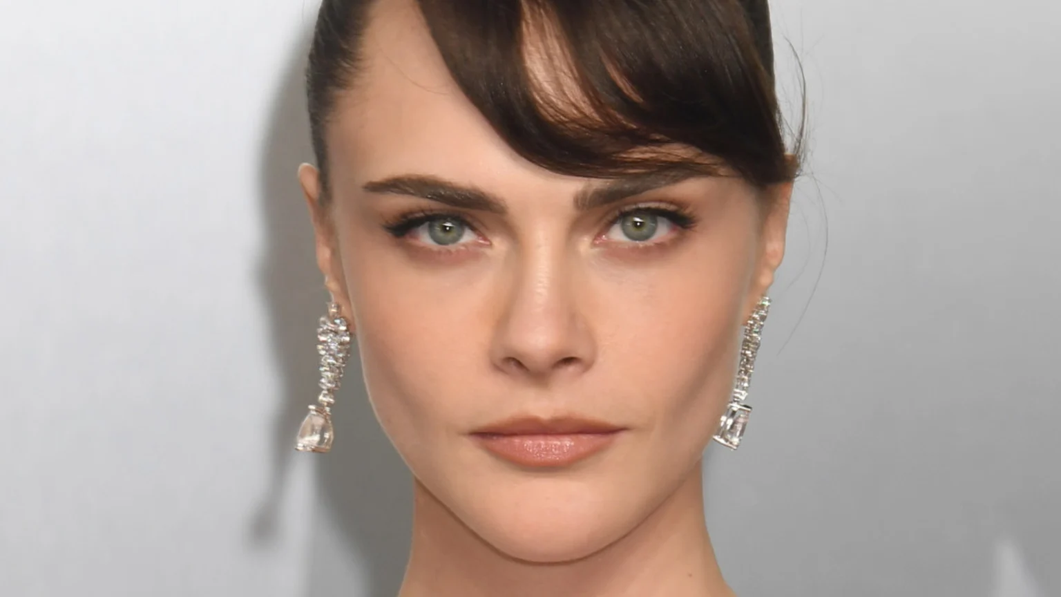 A close-up headshot of Cara Delevingne with dark hair, light blue-green eyes, wearing a subtle makeup look and long, dangling diamond earrings.