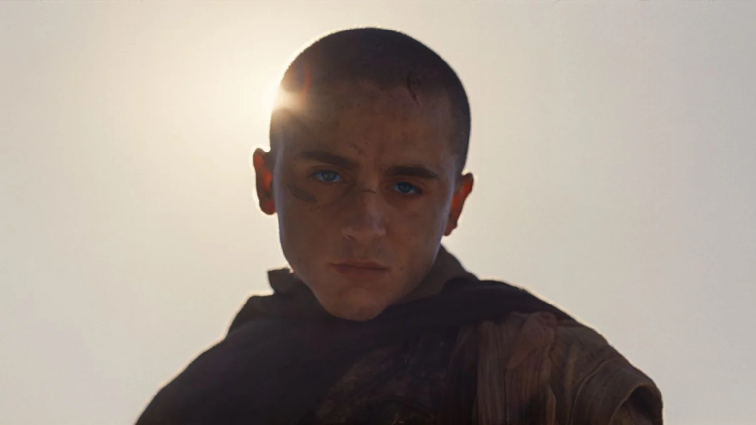 A close-up of Timothée Chalamet as Paul Atreides with bright blue eyes and face scars, looking intensely at the viewer with sun flare behind his head.