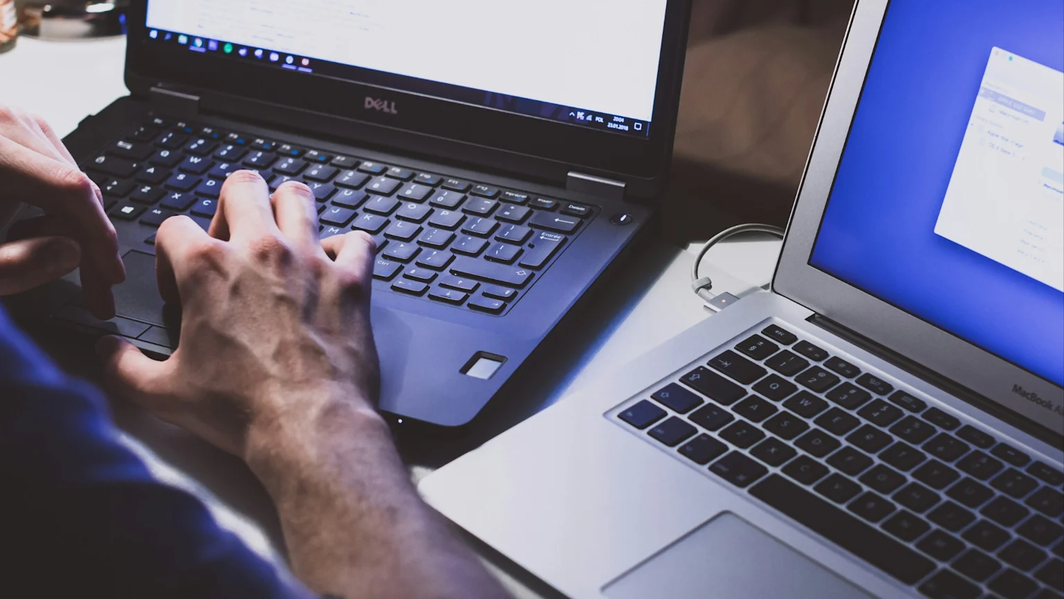 A person types on a Dell laptop displaying a Windows desktop, with a silver MacBook Air also open next to it showing a blue screen, indicating active computing.