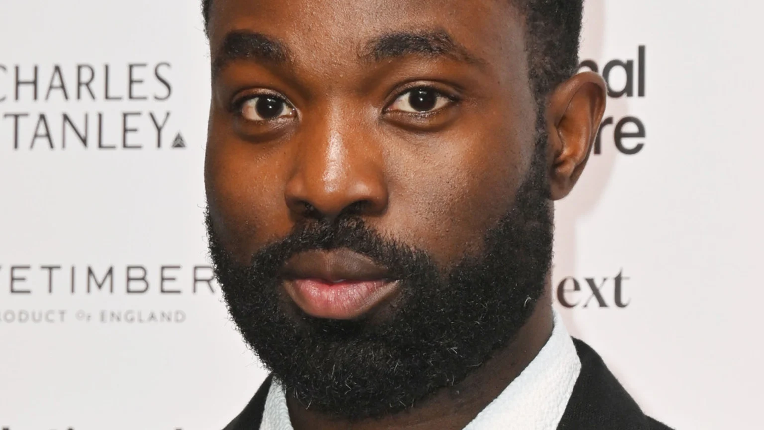 A close-up portrait of a man with dark skin, a dark full beard, and short hair, looking directly forward. He wears a dark jacket over a white shirt, with blurry logos in the background.