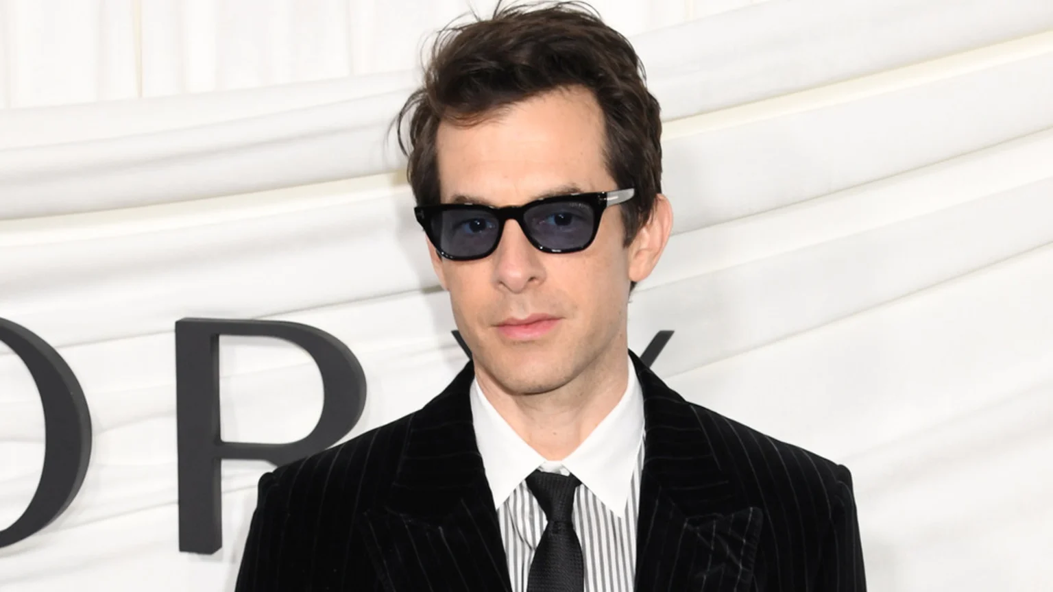 Mark Ronson, wearing a black pinstripe suit, striped shirt, and black tie, looks forward while wearing dark sunglasses against a white background with dark letters.