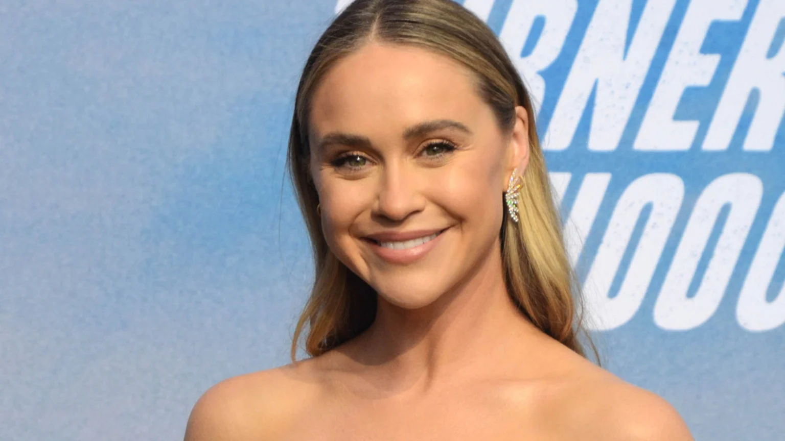 Close-up of a smiling woman with long blonde hair, wearing a strapless top and a sparkly earring, against a light blue background with white text.