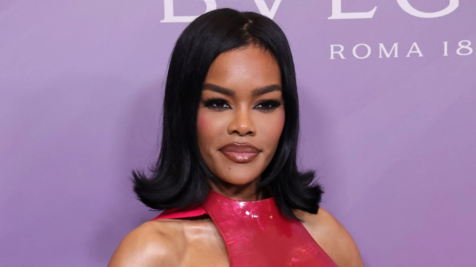 Close-up of a woman with dark skin, a dark bob haircut, and bold makeup, wearing a shiny red top, against a lavender background with partial white lettering.