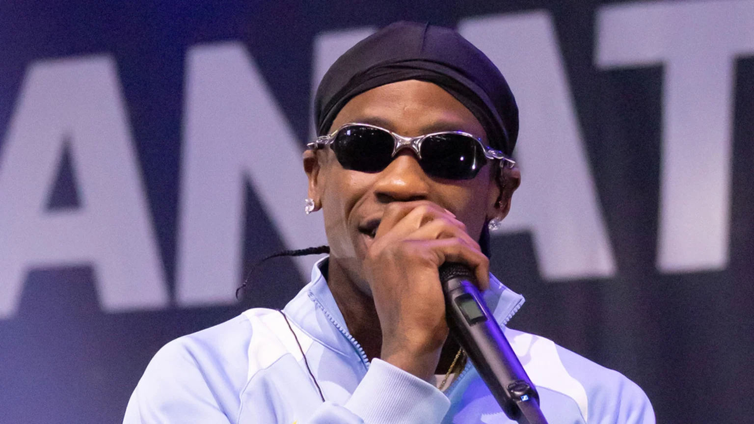 A man in sunglasses, a black durag, and a light blue jacket holds a microphone to his mouth, with blurred white letters in the background.