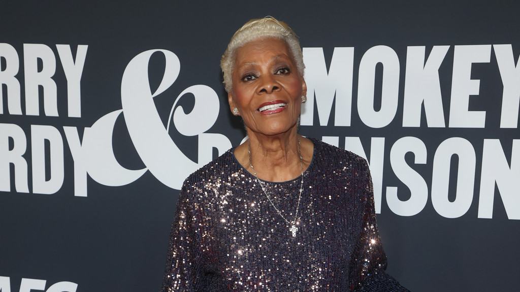 A smiling Dionne Warwick, an older woman with short blonde hair, wears a sparkly top and cross necklace against a dark background with partial white text like "ERRY & MOKEY NSON."
