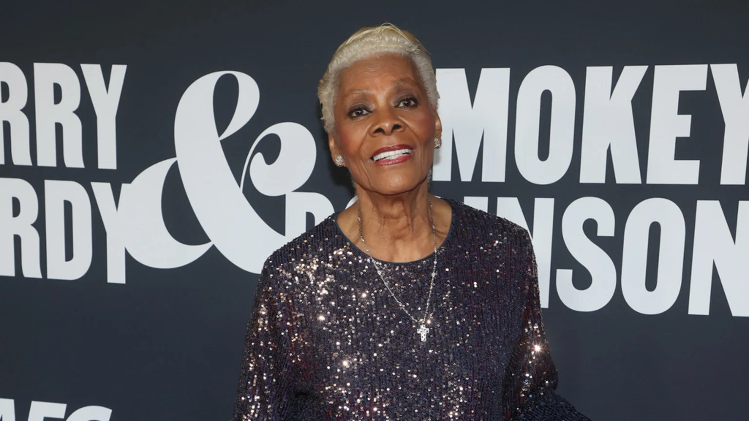 A smiling Dionne Warwick, an older woman with short blonde hair, wears a sparkly top and cross necklace against a dark background with partial white text like "ERRY & MOKEY NSON."
