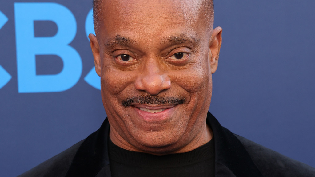 A close-up of actor Rocky Carroll smiling, with a mustache and shaved head, wearing a black jacket against a blue background with a partial light blue "CBS" logo visible on the left.
