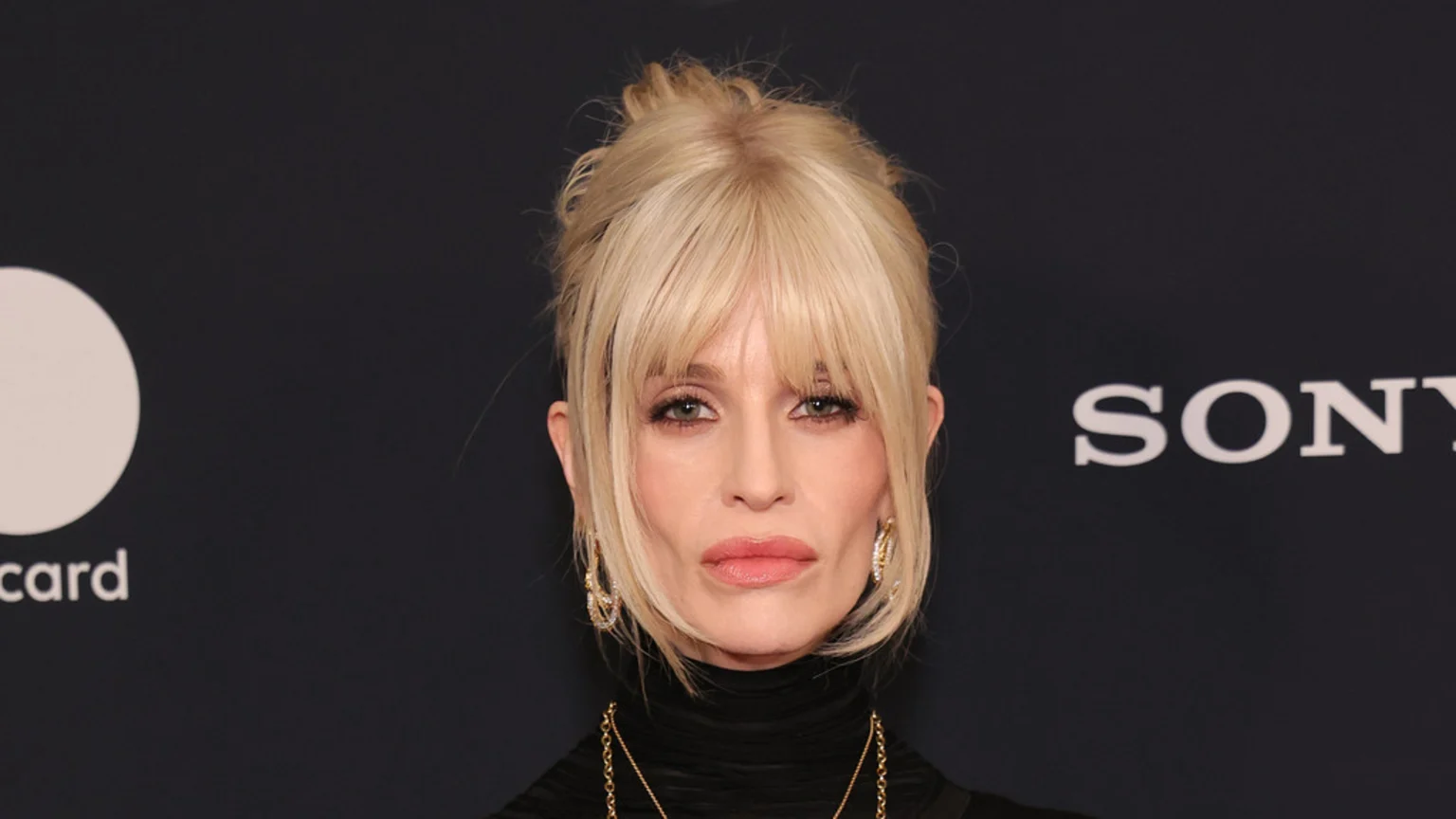 A blonde woman with bangs and a messy bun, wearing gold earrings and a black turtleneck, looks at the camera with a neutral expression, with Mastercard and SONY logos in the dark background.