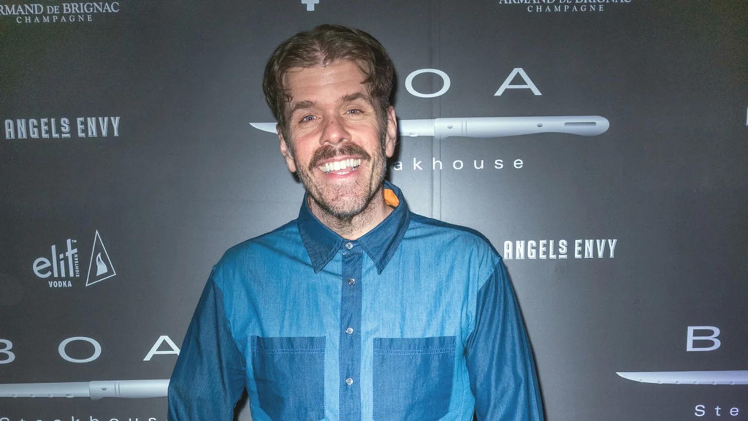 A smiling man with brown hair, a beard, and a mustache, wearing a blue denim shirt, stands in front of a dark grey wall featuring logos for Armand de Brignac, Angels Envy, Elit Vodka, and BOA Steakhouse.