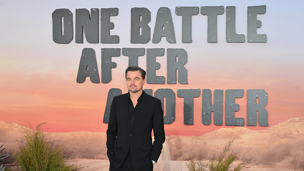 Leonardo DiCaprio in a black suit stands in front of a backdrop displaying "ONE BATTLE AFTER ANOTHER" in grey block letters, against a desert sunset scene.