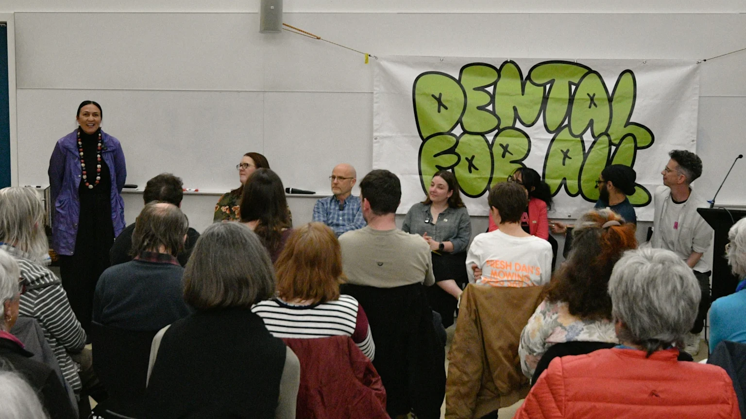 A woman presents at a community event, standing before a green 'DENTAL FOR ALL' banner, a seated panel, and an audience.