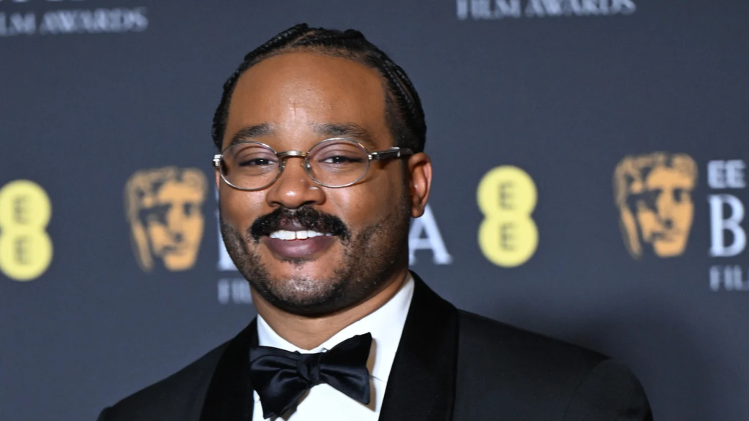 Ryan Coogler, a man with braided hair, a mustache, beard, and round glasses, smiles in a tuxedo against a blurred dark background with yellow and gold award event logos.