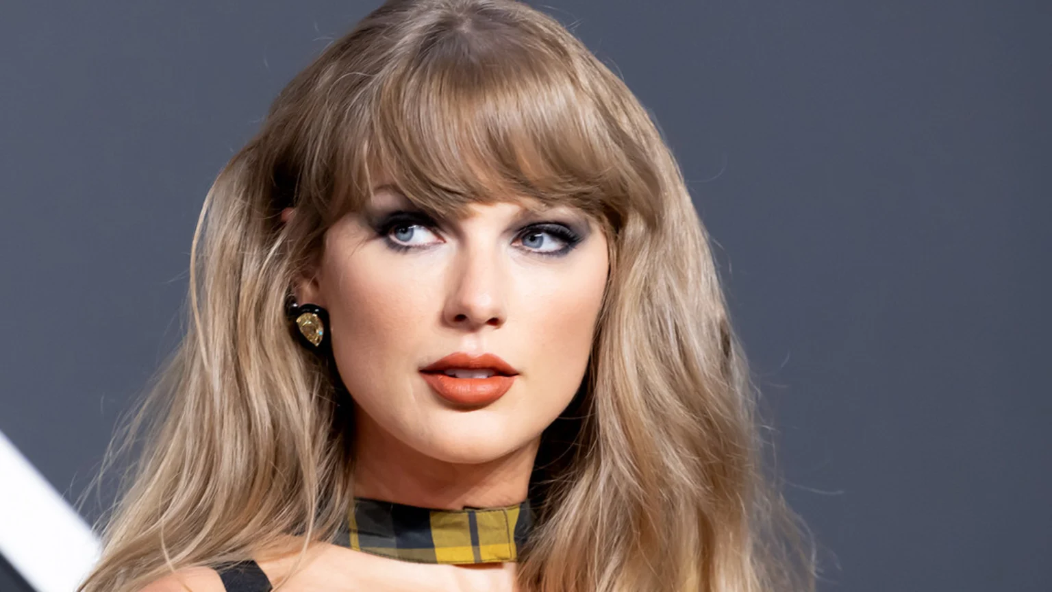 A close-up portrait of Taylor Swift with long wavy blonde hair, dark smoky eye makeup, and rust-colored lipstick. She wears a black and yellow plaid choker, a dark top with a visible strap, and a tear-drop shaped yellow and black earring. The background is solid dark gray with a subtle white diagonal line, photographed indoors under bright artificial lighting in a studio setting.