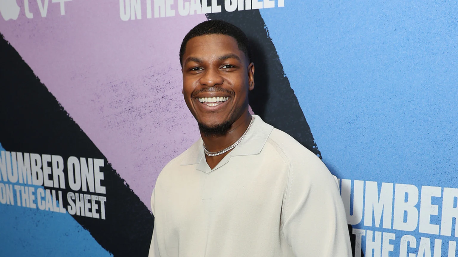 John Boyega smiles broadly, showing his teeth, wearing a light polo shirt and a chain, against a colorful backdrop with text including "APPLE TV+" and "NUMBER ONE ON THE CALL SHEET".