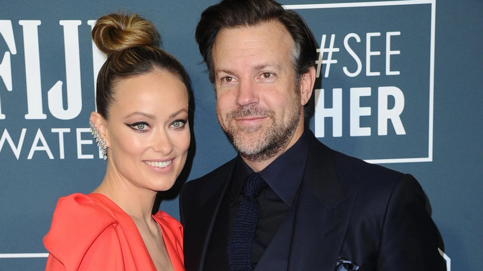 Olivia Wilde, smiling in a red dress with her hair in a bun and an ear cuff, poses next to Jason Sudeikis, who has a beard and wears a dark suit, against a backdrop.
