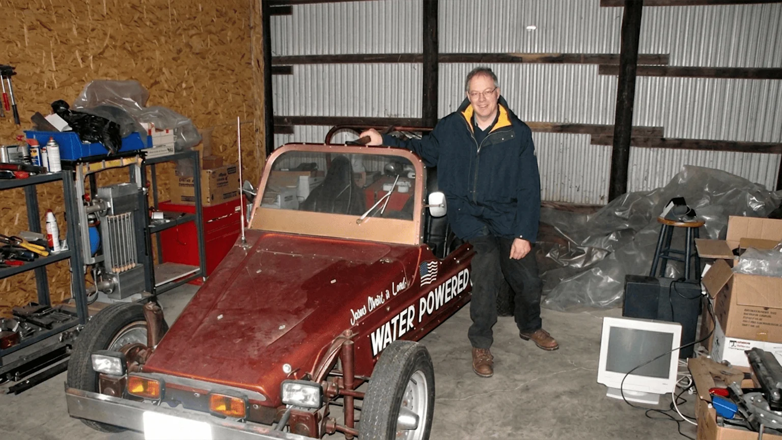A smiling man stands beside a reddish-brown custom car with "WATER POWERED" and an American flag graphic on its side, in a workshop setting.