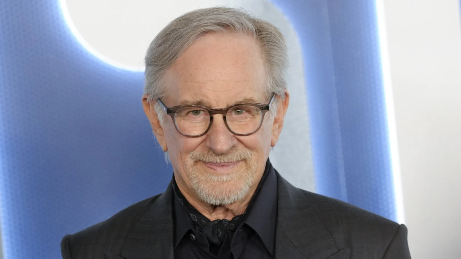 Close-up of Steven Spielberg, a man with grey hair and beard, wearing glasses, a dark suit, and a patterned scarf, smiling at the camera.