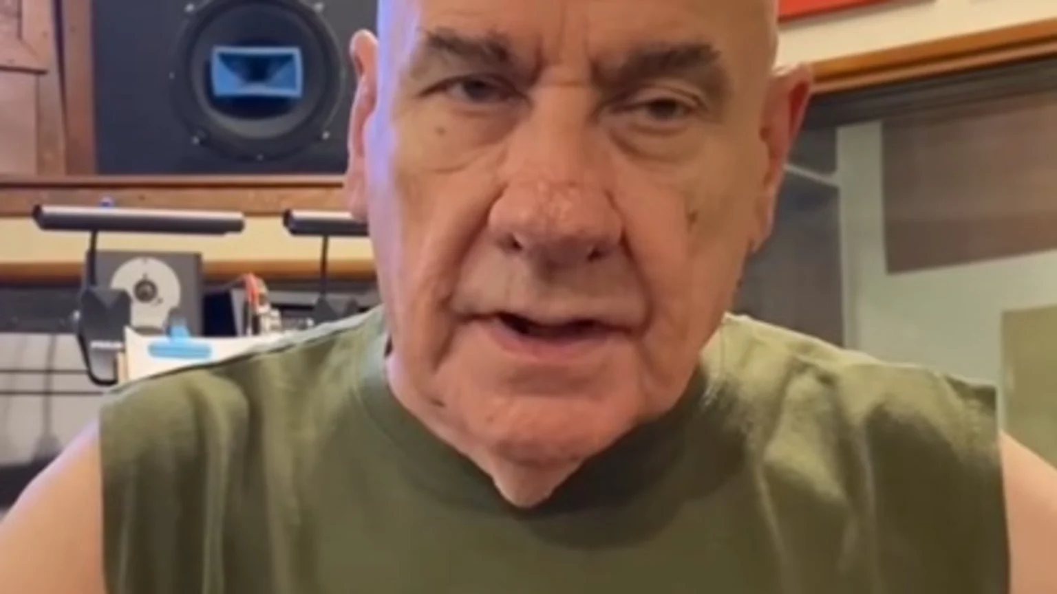 An older bald man in a green sleeveless shirt speaks to the camera from a recording studio, featuring a large built-in speaker unit on the left and red acoustic panels on the right.