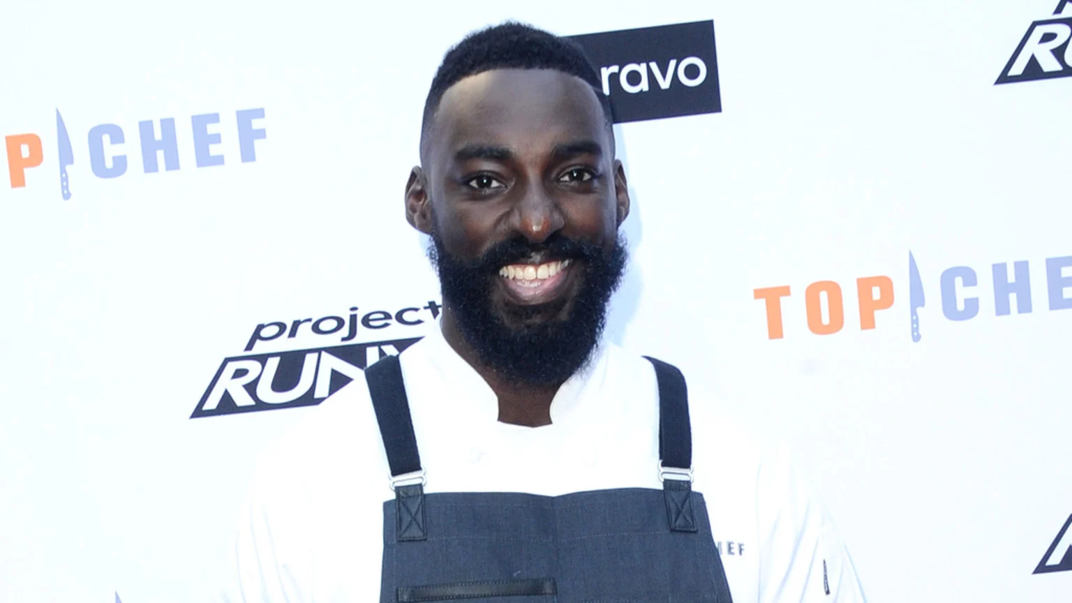 Eric Adjepong, a Black man with a beard, smiles at the camera wearing a white chef's jacket and a dark apron, standing in front of "Top Chef" and "Project Runway" logos.