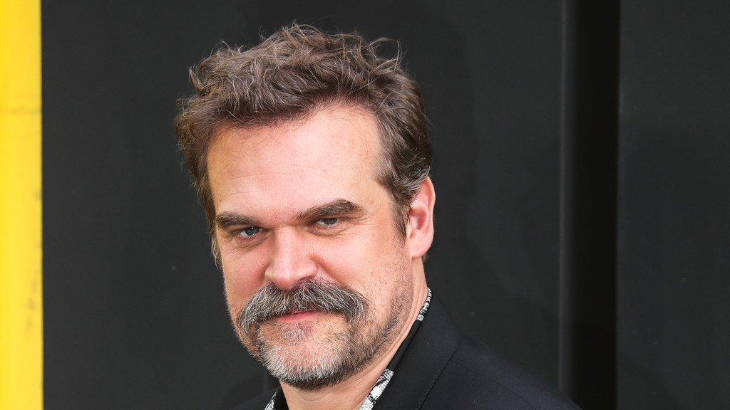 A close-up of actor David Harbour with curly brown hair, a mustache, and a beard, wearing a dark suit and patterned shirt, looking directly at the camera with a serious expression, against a dark background with a yellow stripe.