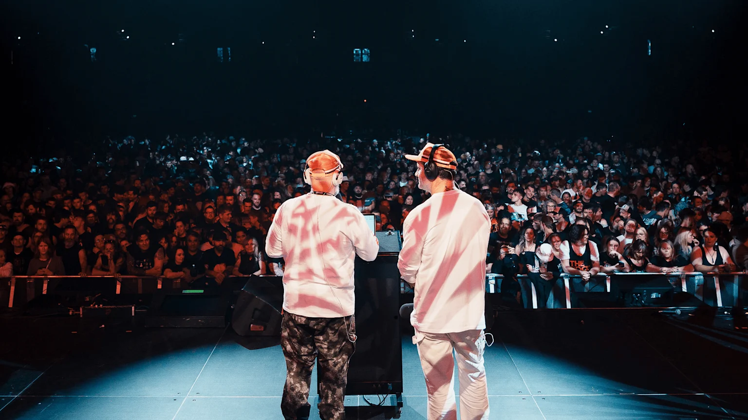 Two performers wearing headphones and hats stand on a stage with their backs to the camera, facing a massive, dimly lit crowd at a music event.