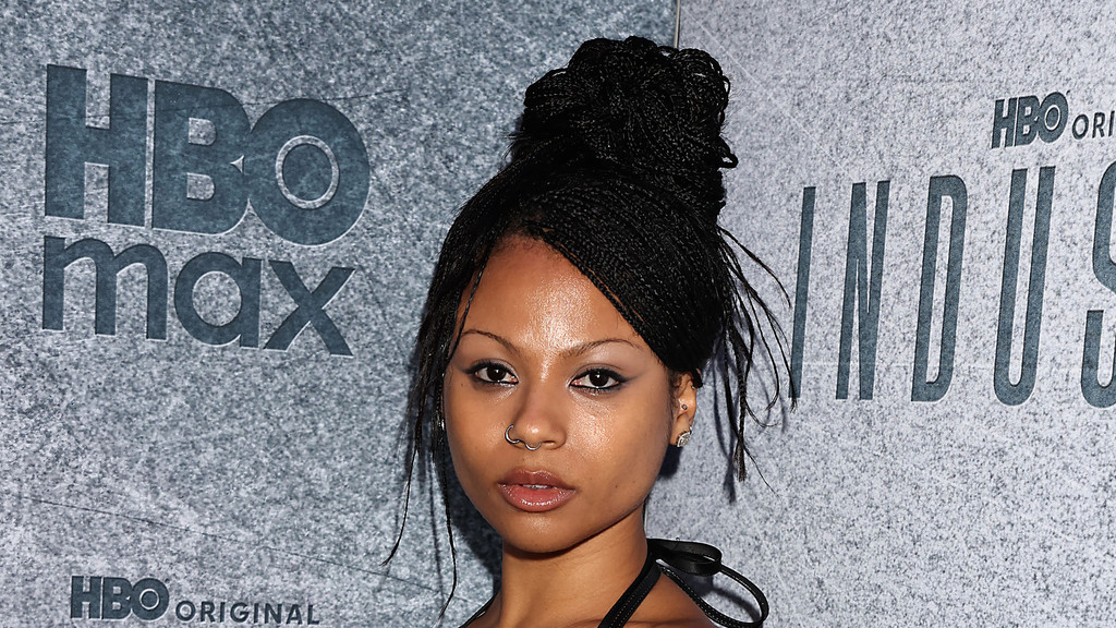 A woman with micro braids styled in a high bun, winged eyeliner, and a nose ring poses in front of a grey wall with "HBO max" and "INDUSTRY" logos.