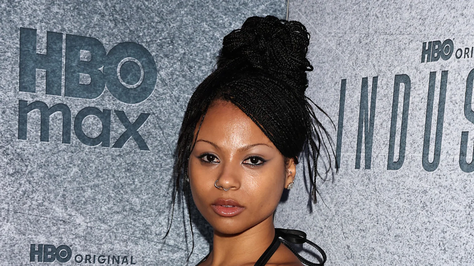 A woman with micro braids styled in a high bun, winged eyeliner, and a nose ring poses in front of a grey wall with "HBO max" and "INDUSTRY" logos.
