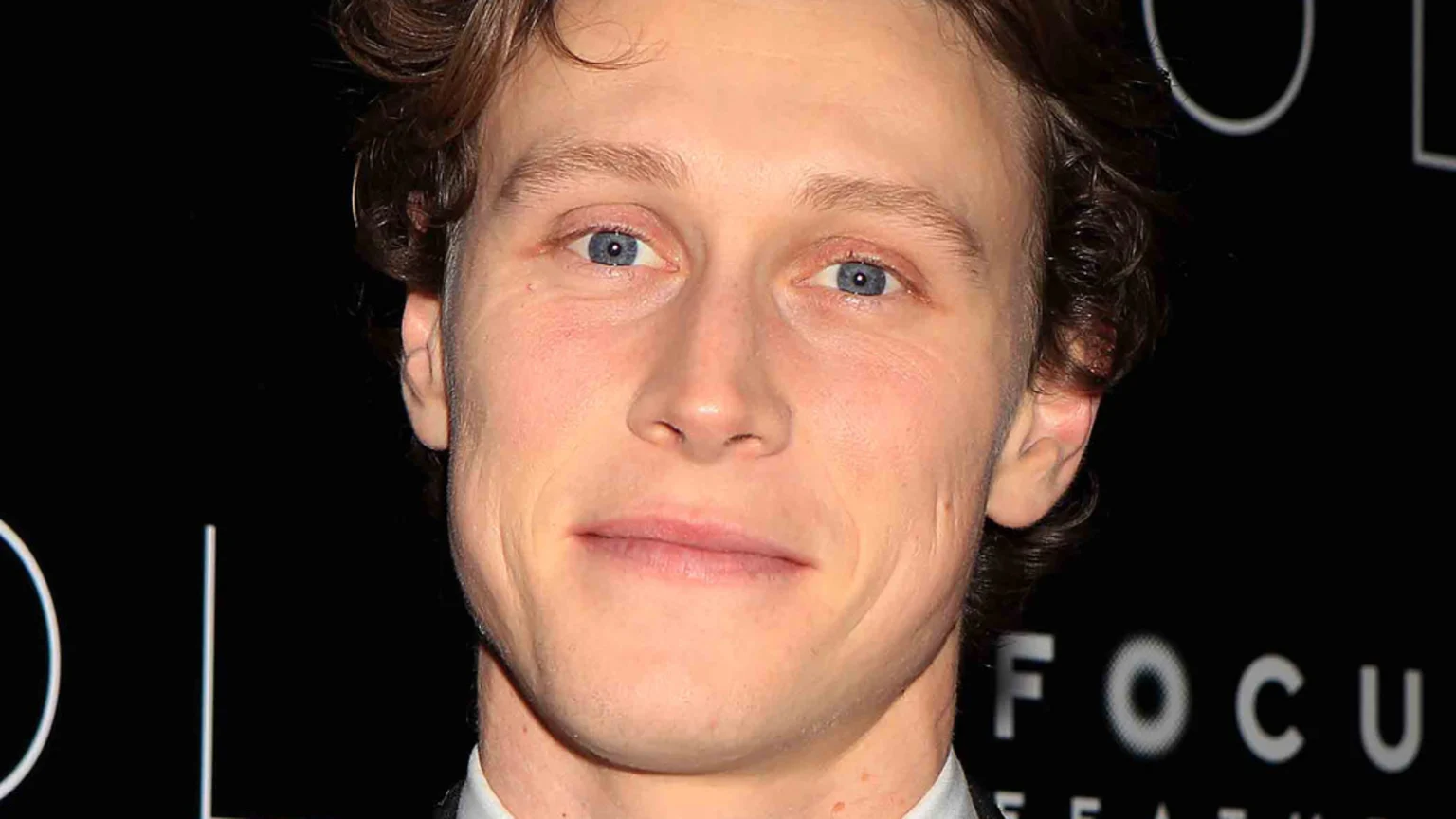 Actor George MacKay with light skin, blue eyes, and wavy brown hair smiles faintly, looking forward, against a dark background with blurred white text.