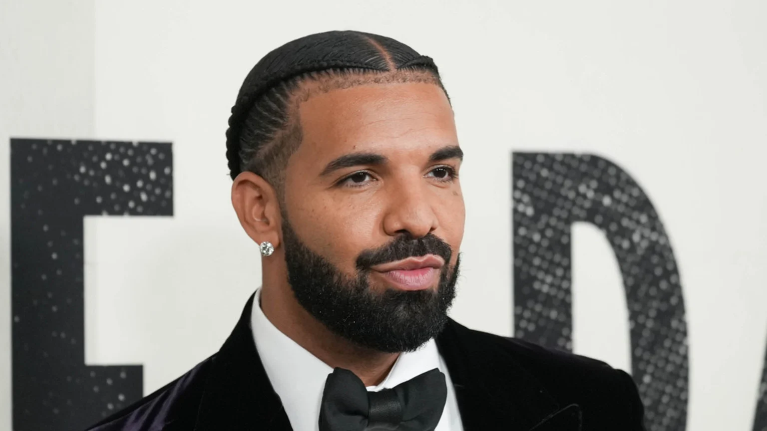 Rapper Drake, in a medium shot, wears a black tuxedo and bow tie, with cornrows, a beard, and a diamond earring, facing right.