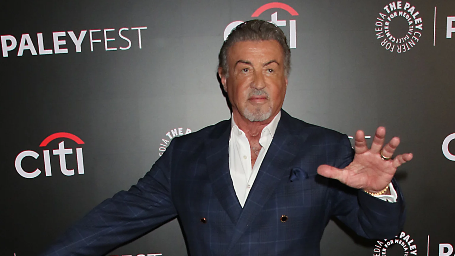 Sylvester Stallone with a beard and graying hair, wearing a blue checkered suit and white shirt, gestures in front of a PaleyFest banner.