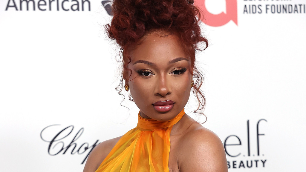 Megan Thee Stallion with reddish-brown curly hair in an updo and an orange halter-neck top looks directly at the camera with noticeable makeup.